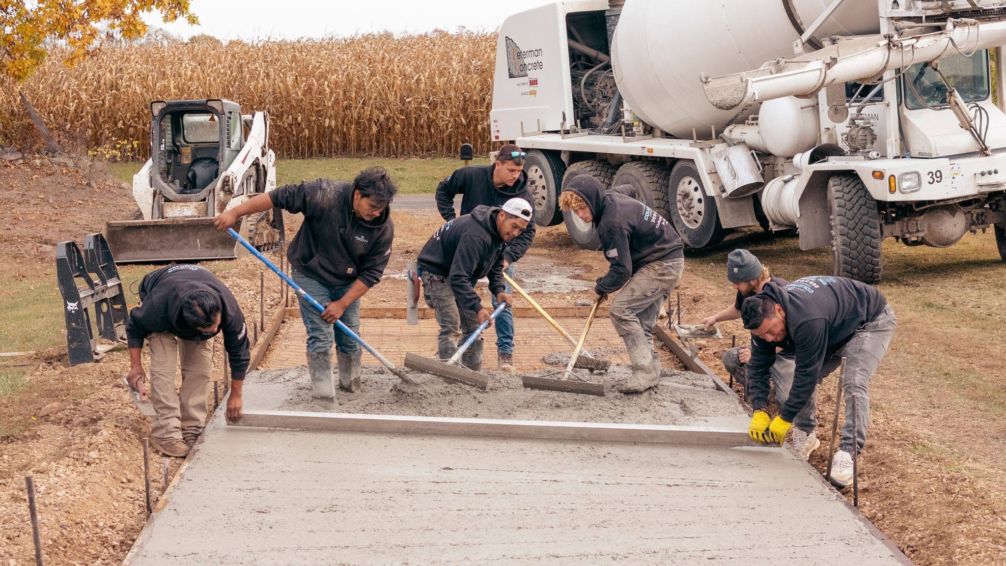Six construction workers, including Chase Courtney, actively spread and level wet concrete on a sidewalk using professional tools. A cement mixer truck and other construction equipment operate in the background, with a cornfield at the edge of the site.
