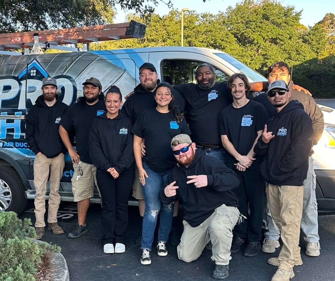 Ten air duct professionals in matching black shirts stand in front of a Pro Shine van with a blue and white logo, outdoors on a sunny day. The team is ready to deliver expert cleaning services for your home.
