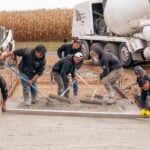 Six construction workers, including Chase Courtney, actively spread and level wet concrete on a sidewalk using professional tools. A cement mixer truck and other construction equipment operate in the background, with a cornfield at the edge of the site.